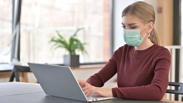 Młoda kobieta z maską twarzy za pomocą laptopa w biurze - Zdjęcie, obraz