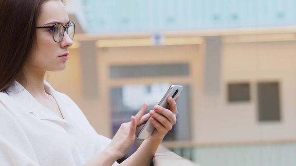 jovem mulher em uma camisa branca e óculos usa smartphone para procurar informações na Internet
. - Foto, Imagem