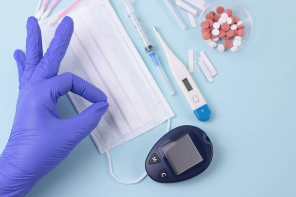 Control of blood sugar levels and prevention of viral infection. Hand in blue latex glove showing OK sign: good test results.Medical background: pills, mask, thermometr, test strips and glucometer. - Foto, imagen