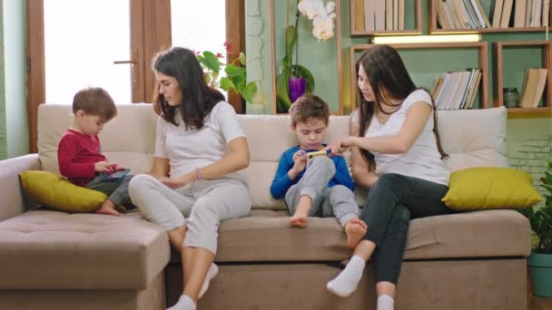 W salonie na kanapie dwie atrakcyjne młode matki z dziećmi spędzają razem czas, podczas gdy chłopcy używają smartfona do gier. - Materiał filmowy, wideo
