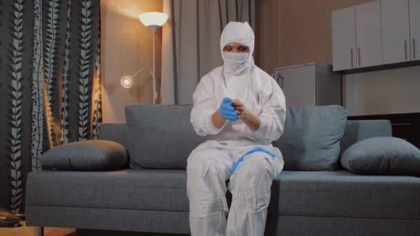 Man wearing protective suit sitting on sofa at home and puts on gloves - Filmmaterial, Video