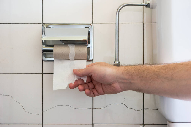 Καυκάσιος αρσενικό χέρι φθάνοντας για ένα σχεδόν άδειο ρολό τουαλέτας δίπλα σε μια μερικώς ορατή λεκάνη τουαλέτας και μεταλλικά υδραυλικά 2020 - Φωτογραφία, εικόνα