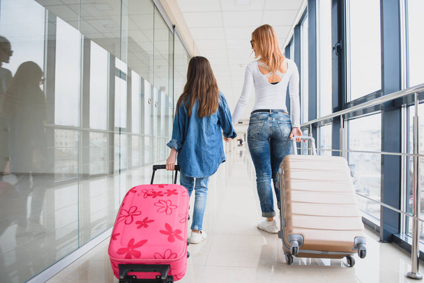 Happy young mother and her daughter walking in the airport terminal while carrying a suitcase. High season and vacation concept. Relax and lifestyles - Foto, afbeelding