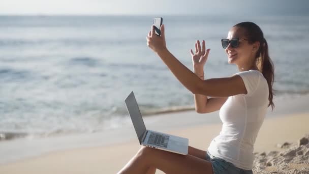 Kobieta z laptopem siedzi na plaży w pobliżu oceanu i używa telefonu do rozmowy wideo - Materiał filmowy, wideo