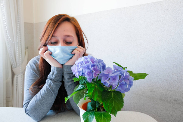 Sadness woman in a medical protective face mask at home quarantine. Girl with blue hydrangea flowers. Pandemic mood. Self-isolation during coronavirus epidemic. Healthy and safety life concept. - Photo, Image