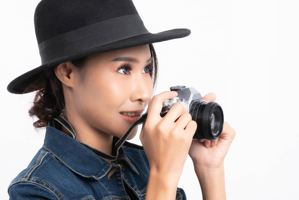 Bella turista donna che indossa un cappello nero e jeans giacca è in piedi per scattare una foto con una fotocamera vintage in felicità in studio girato isolato su sfondo bianco
. - Foto, immagini