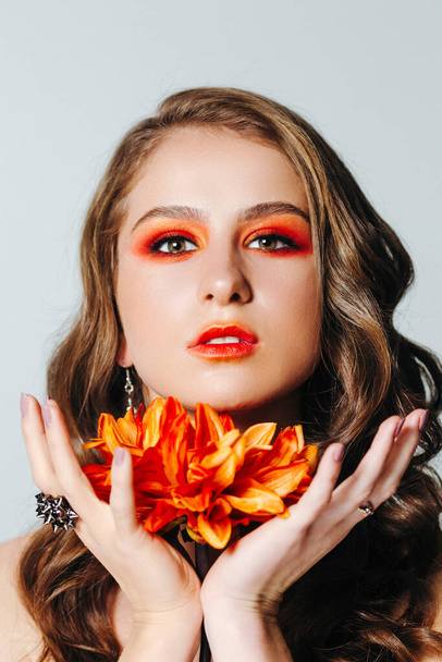 Πορτραίτο ομορφιάς μιας γυναίκας με ένα πορτοκαλί λουλούδι ζέρμπερα και φωτεινό μακιγιάζ. Φθινοπωρινή. Μις Σεπτέμβριος. - Φωτογραφία, εικόνα