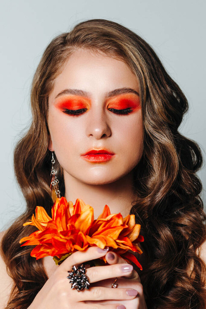 Πορτραίτο ομορφιάς μιας γυναίκας με ένα πορτοκαλί λουλούδι ζέρμπερα και φωτεινό μακιγιάζ. Φθινοπωρινή. Μις Σεπτέμβριος. - Φωτογραφία, εικόνα