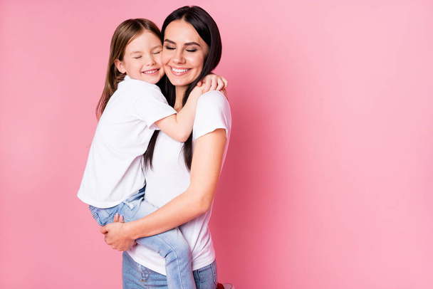 Photo of beautiful young mother hold arms little daughter two ladies hugging best friends lovely feelings eyes closed wear casual t-shirts jeans isolated pastel pink color background - Zdjęcie, obraz