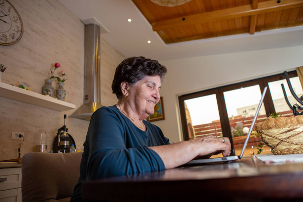 Una donna anziana con i capelli corti neri si siede a un tavolo da pranzo e lavora su un computer portatile. Lei è alla ricerca di alcune nuove ricette di cucina online e lei è premuroso e ridendo
. - Foto, immagini