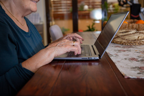 A senior woman with black short hair in a turquoise shirt is typing on a gray laptop keyboard. She laughs and is joyful and is in the dining room at the wooden table. - Photo, Image