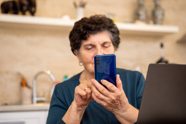 トルコ石のシャツに黒い髪のシニア女性がキッチンに座って、スマートフォンを使用しています。彼女は幸せで笑っている。電話はメタリックブルーで、その前にはノートパソコンがあります。. - 写真・画像