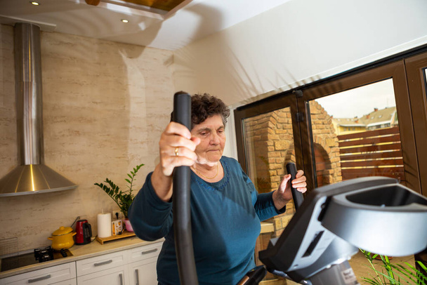 A senior woman with black short hair in a turquoise T-shirt is practicing on a Nordic ski simulator. She is serious and thoughtful because it is not easy for her to train. She's in the kitchen. - Photo, Image