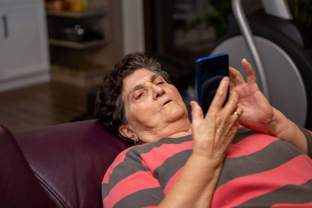 A senior woman lying in her hands has a smartphone as she searches something on the internet. She is a lady of the age but loves modern things modern technology. She's tired and serious. - Foto, Imagen