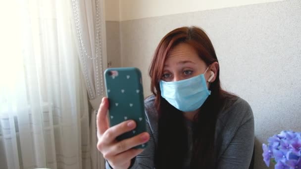 Vrouw met een medisch beschermend gezichtsmasker thuis in quarantaine. Meisje met mobiele telefoon, videogesprek. Pandemische stemming. Vrouw die online spreekt, internet communicatie. Zelfisolatie tijdens de epidemie. - Video