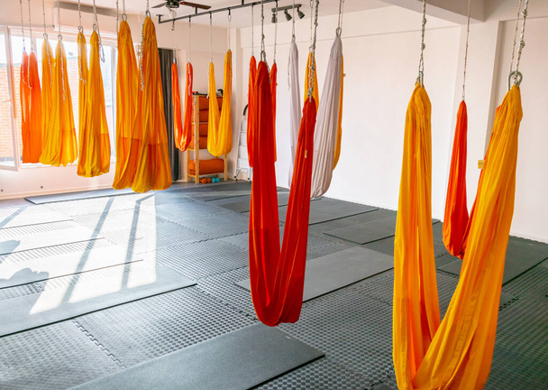 Sala de ioga na luz da manhã com foor preto e paredes brancas, tapetes no chão e vermelho, laranja, redes de ioga penduradas brancas sem pessoas. Aulas de ioga e acessórios de alongamento. 2020
 - Foto, Imagem