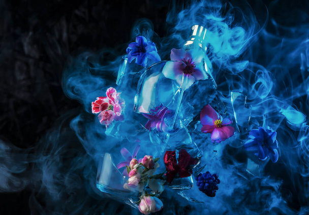 A glass perfume bottle shatters and bright spring flowers and clouds of blue and purple vapor burst out of it against a dark background - Foto, Imagem