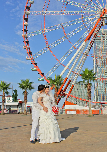 Couple of newlyweds. Bride with a red bouquet in a wedding dress at the palm trees and ferris wheel on background. Happy wedding portrait. Concept marriage, just married, family, honeymoon. - Photo, Image