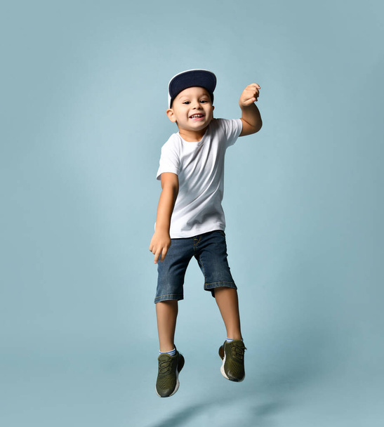 Little brunet child in white t-shirt, denim shorts and khaki sneakers. Jumping up, holding gray cap, posing on blue background - Photo, Image