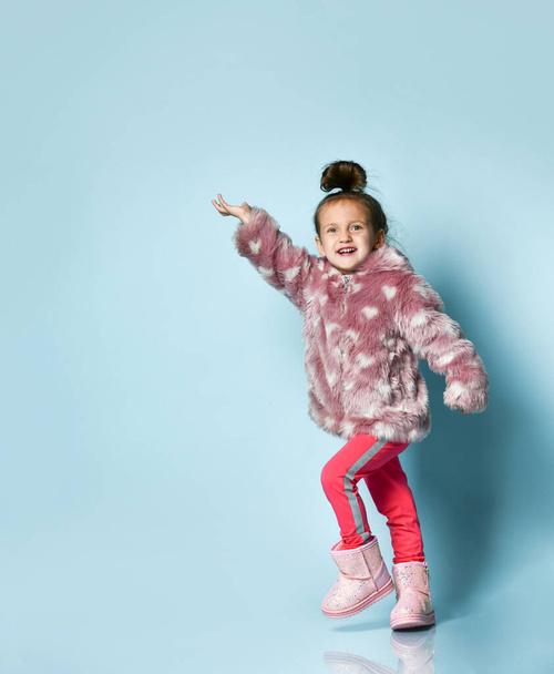 Little girl dressed in pink faux fur coat, pants and boots. She smiling, gesticulating, posing on blue background. Full length - Fotografie, Obrázek