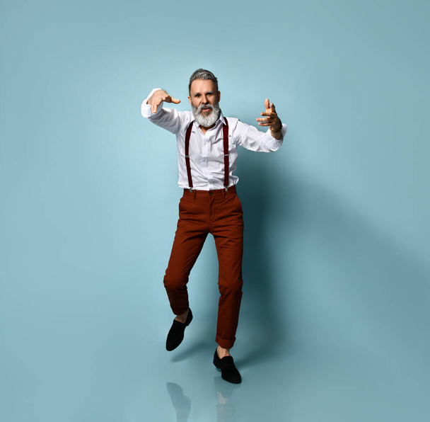 Aged male in white shirt, brown pants and suspenders, black loafers. He is showing thumbs up, walking on blue studio background - Valokuva, kuva