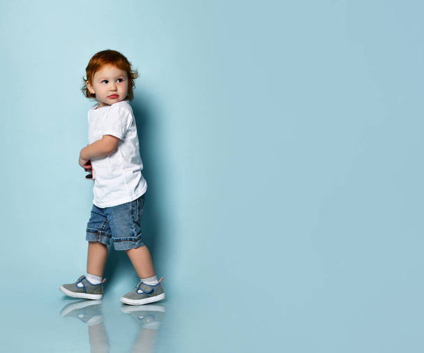 Ginger toddler boy or girl in white t-shirt, socks and shoes, denim shorts. Holding sunglasses, posing sideways on blue background - Photo, Image