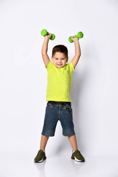 Child in yellow t-shirt, denim shorts, khaki sneakers. Smiling, raised hands up holding green dumbbells, posing isolated on white - 写真・画像