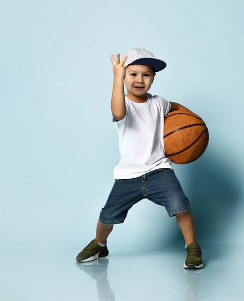 Boy in t-shirt, cap, shorts and sneakers. Holding basketball ball, smiling, showing three fingers, posing on blue background - Φωτογραφία, εικόνα