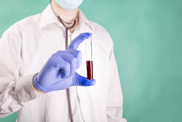 A young doctor, laboratory practitioner with a medical face mask and gloves, holding a test tube with blood sample, on a green background. Periodic blood test. Coronavirus test. A corona pandemic. - Foto, afbeelding