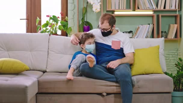 Coronavirus baba kavramı ve küçük oğlu kanepede karantinada. Koruyucu maske takıyorlar. Maske takıp öpüşüyorlar ve akıllı telefondan birlikte vakit geçiriyorlar. - Video, Çekim