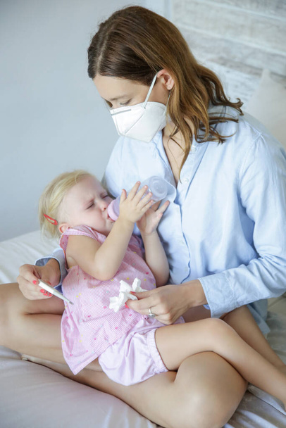 Worried mother sitting on bed and holding her little toddler girl with cold or flu symptoms. Mother wearing protective surgical mask. Coronavirus outbreak and self-quarantining concept. - Photo, Image