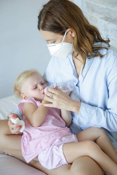 Беспокойная мать сидит на кровати и держит свою маленькую девочку с симптомами простуды или гриппа. Мать носит защитную хирургическую маску. Вспышка коронавируса и концепция самокарантина
. - Фото, изображение