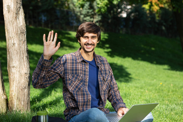 Young brunet freelancer with beard and mustache sitting with a laptop in a park on grass and shake his hand saying hello. Work at any place - Valokuva, kuva