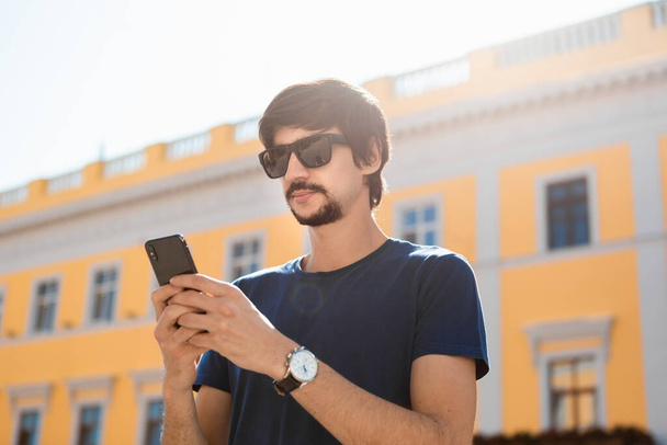 Young brunet man in sunglasses with beard and mustache looking at the screen of a mobile phone walking down the street. Concept of communication - Photo, Image