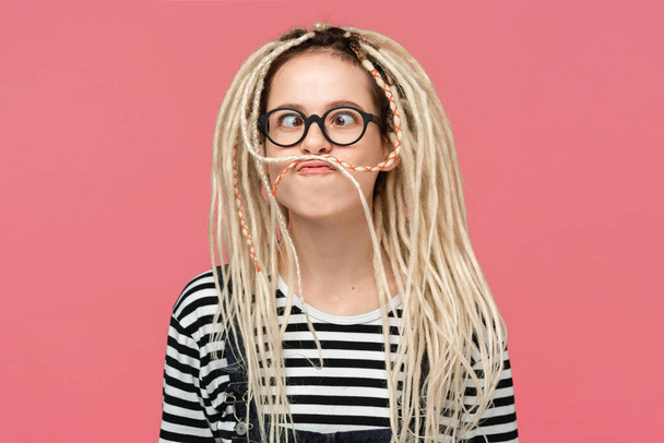Retrato de una joven con gafas vestidas con jersey y camisa a rayas haciendo rostros con sus largas rastas rubias paradas sobre fondo rosa. Concepto de divertirse
 - Foto, imagen