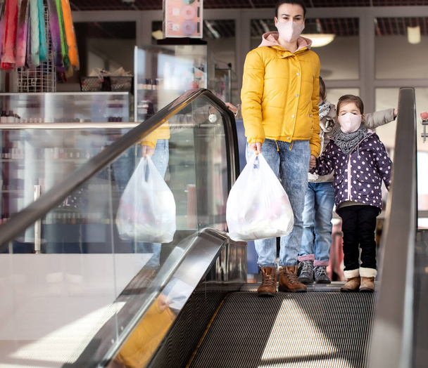 Mutter und Kinder kaufen im Lebensmittelgeschäft ein. Sie gehen die Rolltreppe hinunter. Während der Quarantäne tragen sie Masken. Coronavirus-Pandemie . - Foto, Bild
