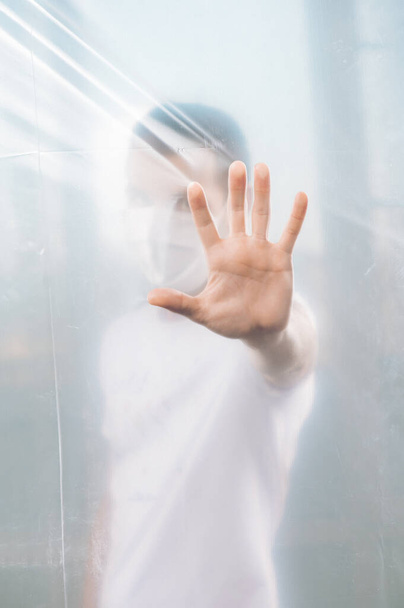 un hombre en cuarentena está de pie en una habitación con la mano apoyada en una barrera de película de plástico
. - Foto, imagen