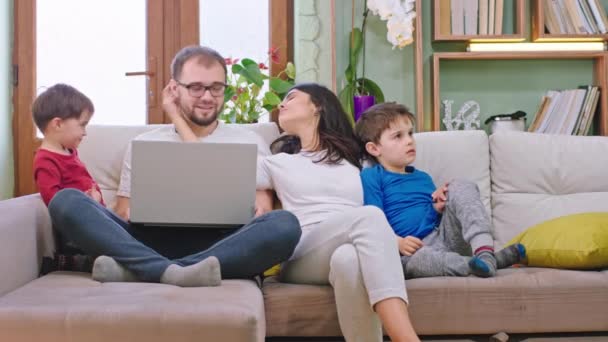 Молода приваблива сім'я на дивані залишається вдома на карантині вони проводять весело час разом граючи в чаті, поки тато працює з дому на ноутбуці
 - Кадри, відео