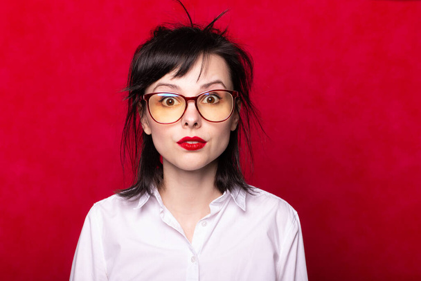 κορίτσι σε λευκό πουκάμισο και γυαλιά με κόκκινα χείλη, κόκκινο φόντο - Φωτογραφία, εικόνα
