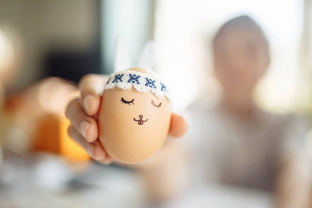 Ένα μικρό πασχαλινό αυγό με ζωγραφισμένο κουνελίσιο πρόσωπο και αυτιά στα χέρια ενός νεαρού έφηβου. Πασχαλινές διακοπές διακόσμηση και προετοιμασία έννοια. Ιερή θρησκευτική ημέρα - Φωτογραφία, εικόνα