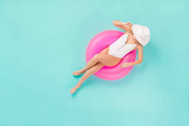 Yüksek açılı, üst açılı, zarif, bronz tenli, yüzme havuzunda oturan bir kadının fotoğrafı. Büyük bir şamandıra. Güneş şapkalı, beyaz yüzme tulumu giyin. - Fotoğraf, Görsel