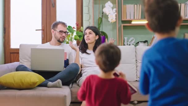 Młoda rodzina z dwójką dzieci w salonie podczas siedzenia na kanapie dwójka małych dzieci biegnie do rodziców z książkami do czytania opowieści - Materiał filmowy, wideo