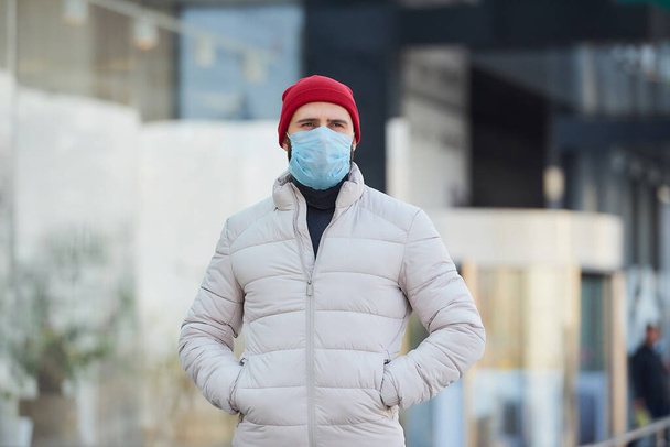 Mężczyzna rasy kaukaskiej noszący medyczną maskę na twarz, aby uniknąć rozprzestrzeniania się koronawirusu (COVID-19). Facet w masce chirurgicznej na twarzy z powodu pandemii przebywającej w centrum miasta.. - Zdjęcie, obraz