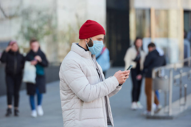 拡散性コロナウイルス(COVID-19)を避けるために医療面マスクを着用している白人男性。町の中心部にスマートフォンを持っているパンデミックのために顔に外科用マスクをした男. - 写真・画像