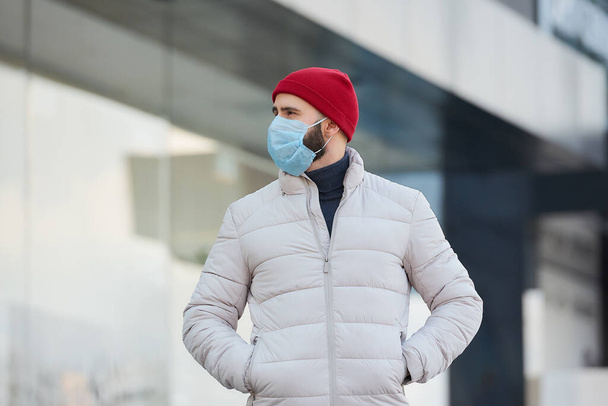 Un homme caucasien portant un masque médical pour éviter la propagation du coronavirus (COVID-19). Un gars avec un masque chirurgical sur le visage à cause de la pandémie qui attend dans la rue
.  - Photo, image