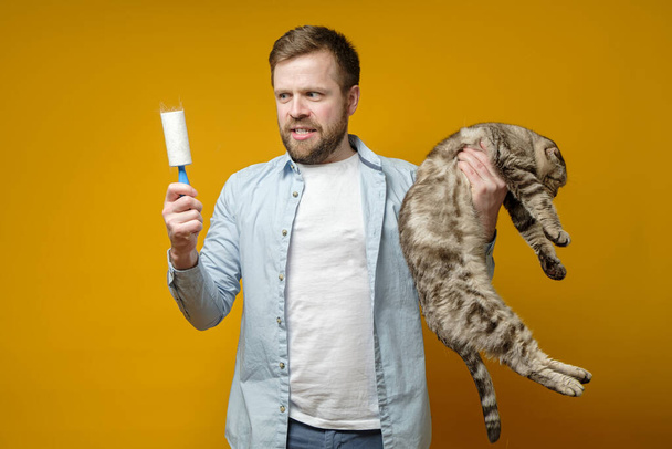 Disgruntled man holds cat in one hand and in the other a roller with adhesive tape in animal hair, which he removed from clothes. - Photo, Image
