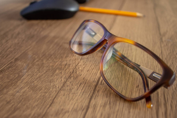 Closeup on a par of glasses with a pencil and mouse on the background, on top of a wooden table - Фото, изображение