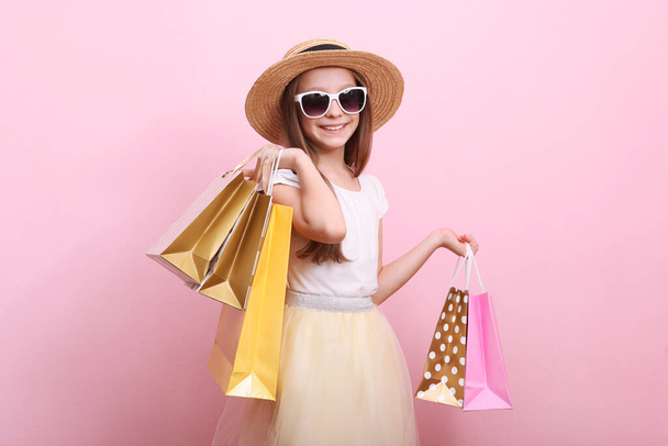 Όμορφο χαμογελαστό κοριτσάκι με τσάντες για ψώνια σε έγχρωμο φόντο. Η έννοια των παιδικών αγορών. - Φωτογραφία, εικόνα