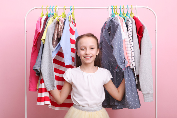 Aranyos vidám kislány választja ruhák padló akasztók. Gyerekruhák, gyerekvásárlás. Divatos ruhák gyerekeknek. - Fotó, kép