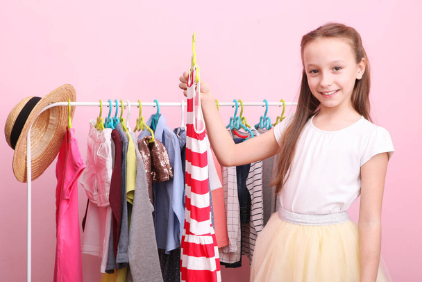 Roztomilá veselá holčička si vybírá oblečení s podlahovými ramínky. Dětské oblečení, dětské nákupy. Módní oblečení pro děti. - Fotografie, Obrázek